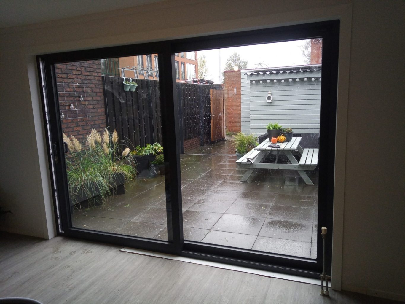 Uitzicht op een betegelde achtertuin met planten en een houten tafel.